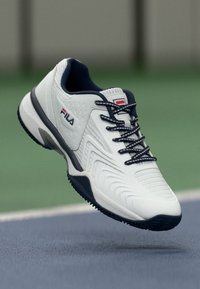 Fehér sportcipő texturált felszínen, tengerészkék részletekkel és fekete gumitalppal; fűzőkkel és márka logóval az oldalon.