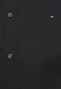 Czarna koszula z guzikami, wykonana z gładkiego materiału, z szarymi plastikowymi guzikami i małym haftowanym logo w kolorach czerwonym, białym i niebieskim.