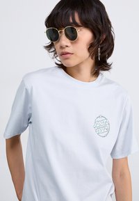 T-shirt di cotone azzurro chiaro con maniche corte, caratterizzato da un logo circolare "Santa Cruz" sul petto in un design colorato.