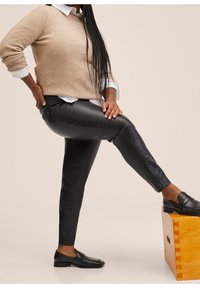 Černé legíny z umělé kůže s elegantním designem, spárované s béžovým svetrém a černými mokasíny, na neutrálním pozadí.