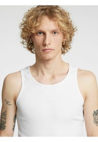 Hvit bomulls singlet med ribbet tekstur og brede skulderstropper. Modellen har krøllete blondt hår og synlige tatoveringer på armene.