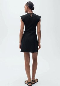 Robe noire courte avec des manches courtes et un détail en forme de trou de serrure à l'arrière, présentant une silhouette ajustée et une texture lisse. Sandales avec des clous.