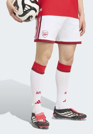 Osoba nosząca krótkie spodenki piłkarskie Arsenalu, białe skarpetki z czerwonym obszyciem i czarne korki, trzymająca czarno-białą piłkę nożną Adidas.
