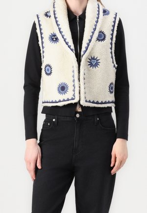 Haut à manches longues et pantalon noirs portés sous une veste sans manches en polaire blanche, brodée de motifs bleus de soleil et de cœur le long des bords et à l'avant.