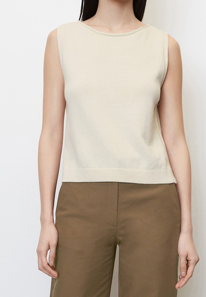 Donna che indossa un top senza maniche di maglina color avorio e pantaloni marroni a vita alta, in piedi contro uno sfondo chiaro e semplice.