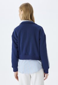 Jersey azul marino con un diseño corto, puños acanalados y una camiseta rayada blanca asomando por debajo. Tejido suave con líneas limpias.