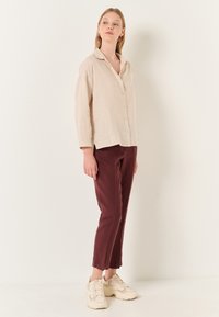 Camicia in lino beige chiaro con bottoni e colletto, abbinata a pantaloni borgogna e sneakers chiare, mostrata di lato su uno sfondo neutro.