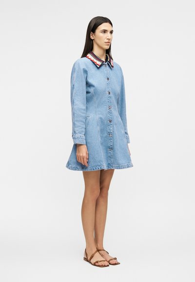 Γυναίκα όρθια, φοράει ένα ανοιχτό μπλε φόρεμα από τζιν με κουμπιά και πολύχρωμο κολάρο με λουλούδια, καθώς και καφέ επίπεδα σανδάλια, σε απλό φόντο.