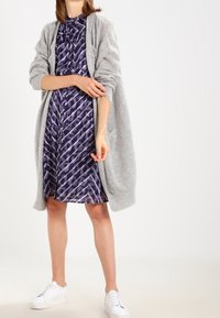 Modelka v květinových fialových šatech s šedým svetrem, bílými teniskami a uvolněným postojem na světlém pozadí.