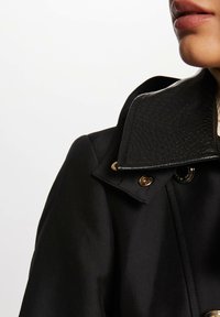 Svart trench coat med en teksturert svart krage, gullfargede detaljer, og en glatt stofffinish. Nærbilde av kragen og stoffet.