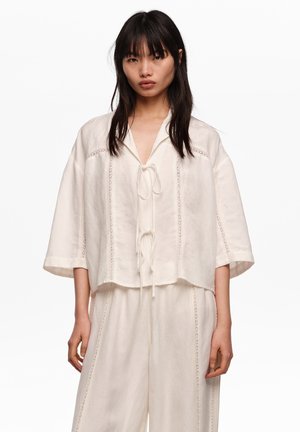 Mujer con conjunto suelto de lino color crema con mangas medias y costuras verticales decorativas, de pie frente a un fondo liso.