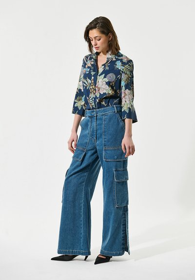 Femme debout, la tête légèrement inclinée, portant un chemisier à motifs floraux et un jean cargo large avec des chaussures noires pointues.