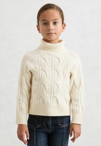 Bjelkasti pulover s visokim ovratnikom i pletenim uzorkom, s rebrastim manžetama i rubom, u kombinaciji s tamnoplavim trapericama koje imaju vidljive džepove.