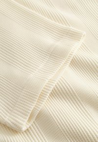 Krém színű, bordázott anyag közeli képe, látható varrással és hajtott széllel, amely kiemeli a textúrát és a kötési mintázatot.