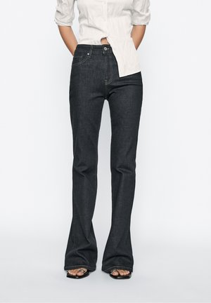Femme portant un jean noir flare taille haute associé à une chemise blanche à boutons et des sandales noires à talons ouverts.