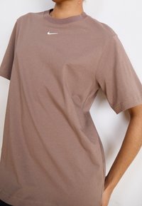 Person med en brun oversized Nike-t-shirt med en liten vit Nike-logotyp på bröstet, armen böjd vid midjan, vit bakgrund.