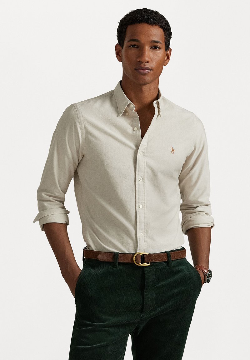 Chemise à col boutonné en tissu beige clair avec une texture subtile, associée à un pantalon en velours côtelé vert foncé et une ceinture marron avec une boucle en or.