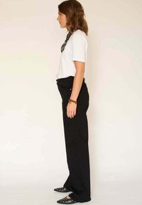 Pantalon noir taille haute, coupe décontractée, associé à un t-shirt blanc. Un foulard noir à motifs autour du cou et des chaussures noires pointues cloutées.