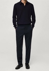 Mężczyzna ubrany w czarną koszulę z długim rękawem i kołnierzykiem, ciemne dopasowane spodnie oraz czarne eleganckie buty, stojący z rękoma w kieszeniach.