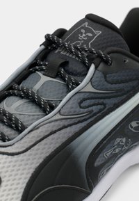 Gros plan sur une chaussure de sport noire et grise avec des lacets à motifs, un graphique de chat souriant sur la languette et des visages de chat sur le panneau latéral.