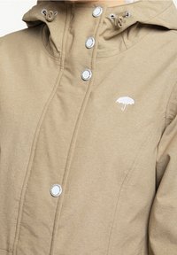 Veste à capuche beige en matière douce. Elle comprend des boutons-pression argentés et un logo brodé d'un parapluie blanc sur la poitrine.