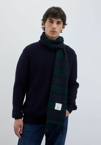 Sciarpa di lana in un design a quadri blu scuro e verde, con estremità frangiate e un'etichetta con logo. Indossata con un maglione navy a trama.