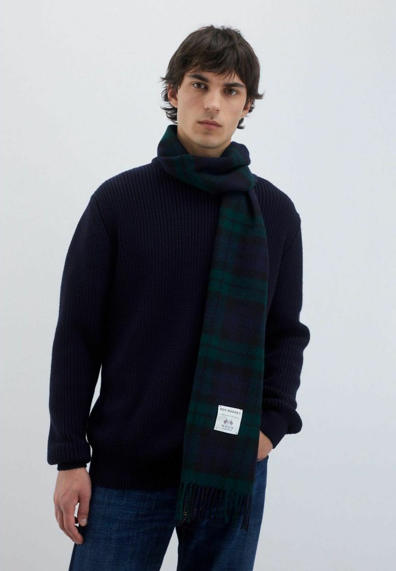 Sciarpa di lana in un design a quadri blu scuro e verde, con estremità frangiate e un'etichetta con logo. Indossata con un maglione navy a trama.