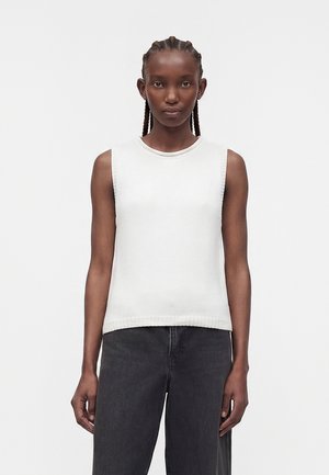 Kobieta z zaplecionymi włosami, ubrana w białą bezrękawnikową prążkowaną bluzkę i czarne spodnie, stojąca na jednolitym jasnym tle.