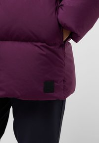 Paarse pufferjack met een gladde textuur, volumineuze secties en een zwart merklabel nabij de zoom. Gemaakt over donkere broeken.
