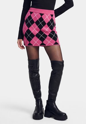 Donna che indossa una minigonna con motivo argyle rosa e nero, calze nere trasparenti e stivali neri sopra il ginocchio con tacco spesso.