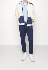 Fleecejacka i cream med marinblå accenter, matchad med en ljusblå zip-up skjorta och marinblå byxor. Vita sneakers fulländar outfiten.