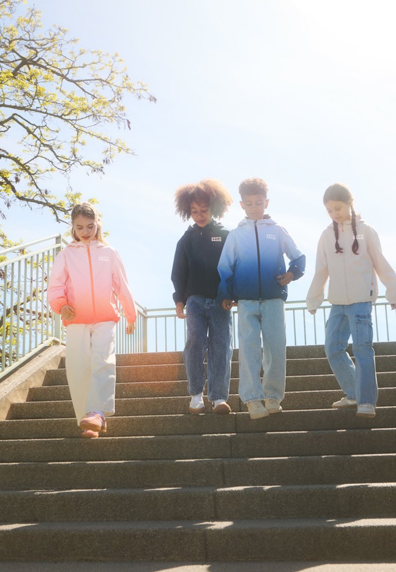 Quatre enfants descendent des escaliers. Ils portent des vestes légères et colorées : dégradé rose, marine, dégradé bleu et gris clair, accompagnées de jeans décontractés.