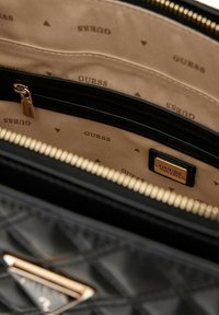 La borsa a tracolla in pelle nera trapuntata presenta un interno rivestito in tessuto beige con il logo "GUESS", una cerniera dorata e un compartimento con zip.