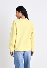 Sweatshirt amarelo com decote redondo, mangas raglan longas, punhos e bainha canelados. Combinado com jeans azul claro, textura de tecido suave.