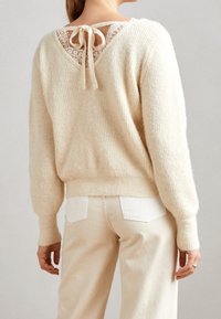 Beige gebreide trui met een V-vormige kant achter met een strik, geribbelde textuur en een relaxte pasvorm, gecombineerd met lichtgekleurde jeans.