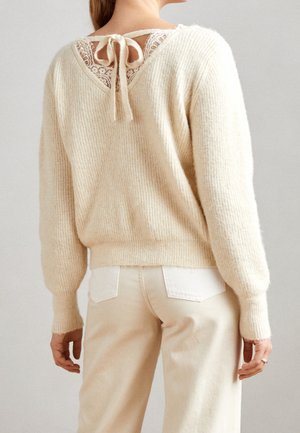 Pull en tricot beige avec un dos en dentelle en forme de V agrémenté d'un nœud, texture côtelée et coupe décontractée, assorti à un jean clair.