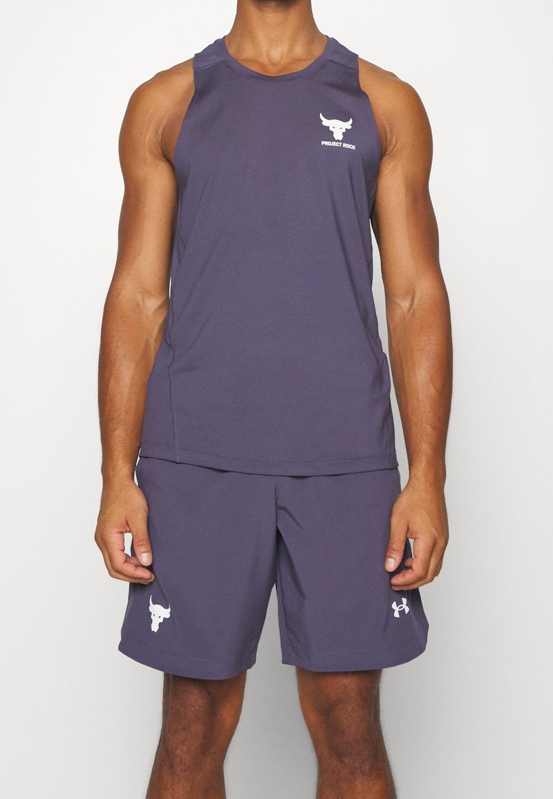 Svalnatý člověk v bezrukávovém fialovém sportovním tričku a odpovídajících šortkách s bílými logy Project Rock a Under Armour.