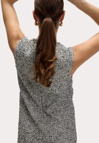 Svart och vit, texturerad ärmlös topp med öppen rygg, featuring en lös passform och ett vävt mönster. Örhängesdetalj synlig.