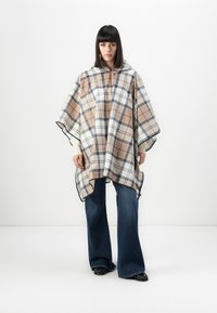 Woman wearing beige, navy, and cream plaid poncho with wide-leg jeans and black shoes, standing against white background.