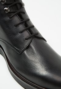 Černá kožená bota s hladkým povrchem, šněrovací design a stříbrné očka. Obsahuje zaoblenou špičku a kontrastní švy.