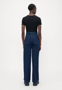 Personne avec des cheveux tressés portant des boucles d'oreilles en argent, un haut noir à manches courtes, un jean évasé taille haute bleu foncé, et des chaussures noires à semelles épaisses.