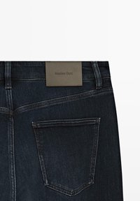 Jeans in denim blu scuro con una striscia rettangolare in pelle marrone etichettata "Massimo Dutti" sulla cintura, caratterizzati da una tasca posteriori con un contorno ricamato.