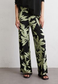 Wide-leg trousers made of smooth fabric, featuring a black base with large green floral patterns. Worn with flat black sandals.