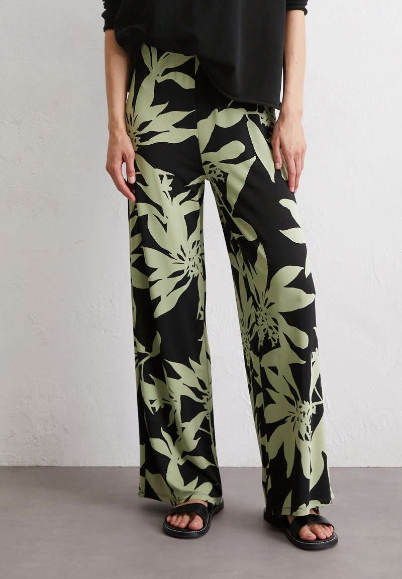 Weite Hose aus glattem Stoff, mit schwarzem Grund und großen grünen Blumenmustern. Tragen mit flachen schwarzen Sandalen.