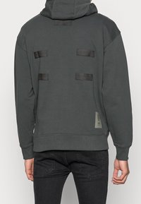 Felpa con cappuccio grigio scuro con tasca a marsupio, cappuccio strutturato e quattro cinghie orizzontali in nylon sulla schiena. Polsini e orlo a costine.