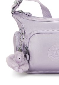 Lilla metallisk crossover taske med en tekstureret overflade, der har en forlomma, sølv hardware og en fluffy abe nøglering som tilbehør.