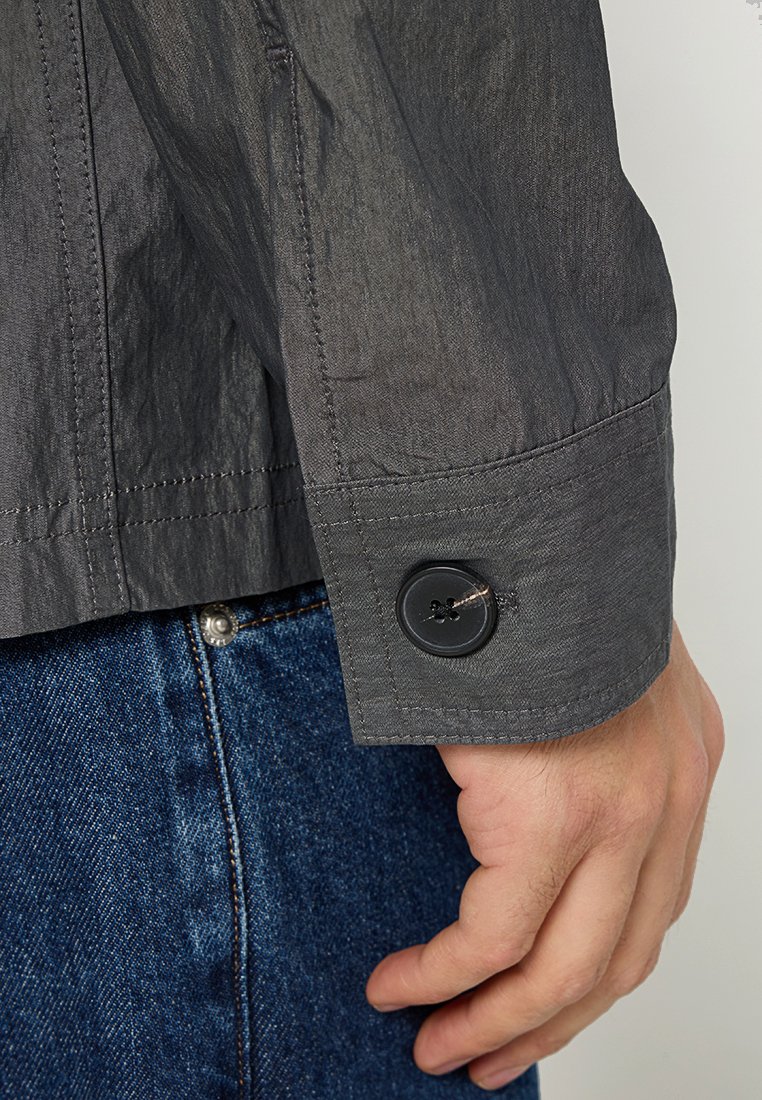 Szary rękaw marynarki zapinanej na guziki z czarnym detalem. Posiada fakturowany materiał i widoczne przeszycia, w połączeniu z deninowym pasem poniżej.