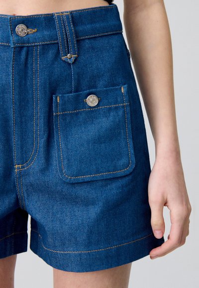 Pantalones cortos de mezclilla en azul profundo, con costuras doradas en contraste, un cierre de botón y un bolsillo frontal con un detalle de broche de metal.