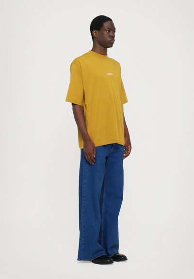 Tricou galben cu mânecă scurtă, având un mic logo roșu, asortat cu blugi dark blue cu croi larg și pantofi negri, pe un fundal simplu.