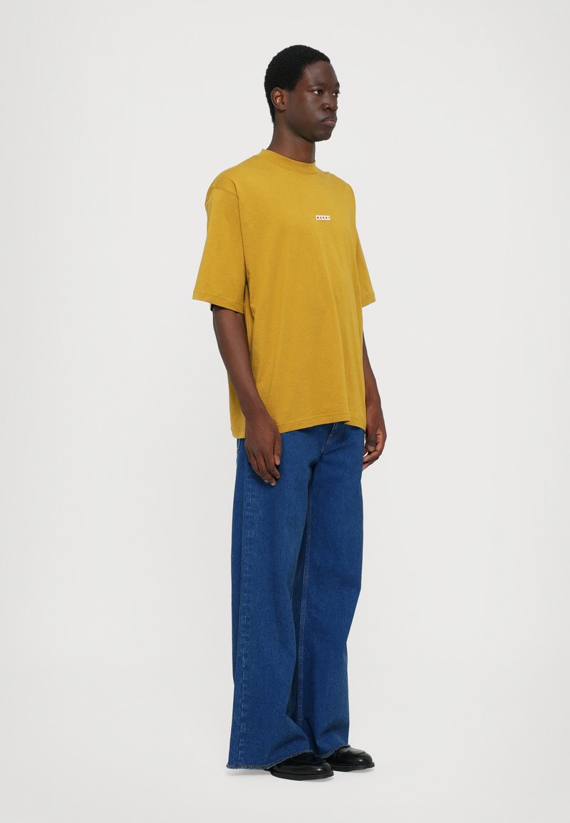 Yellow short-sleeved T-shirt with a small red logo, paired with wide-leg dark blue denim jeans and black shoes, against a plain background.